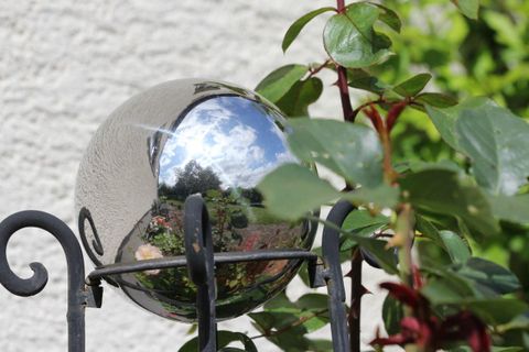 Eine spiegelnde Kugel sitzt auf einer Metallhalterung, umgeben von grünen Pflanzen. Der Himmel und Wolken spiegeln sich in der Kugel wider.