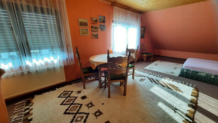 Ein gemütliches Zimmer mit orangefarbenen Wänden und einem runden Tisch. Es gibt Stühle, einen Teppich und viel natürliches Licht von den Fenstern.
