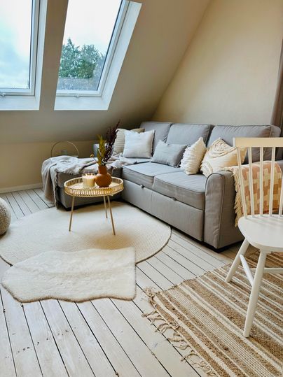 Ein gemütliches Wohnzimmer mit einem grauen Sofa und großen Fenstern. Der Boden ist aus Holz und es liegt ein weicher Teppich im Raum.