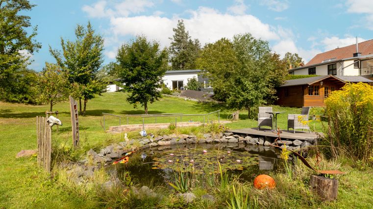 Ein schöner Garten mit einem kleinen Teich und Bäumen. Im Hintergrund sind ein Haus und eine Gartenlaube zu sehen.