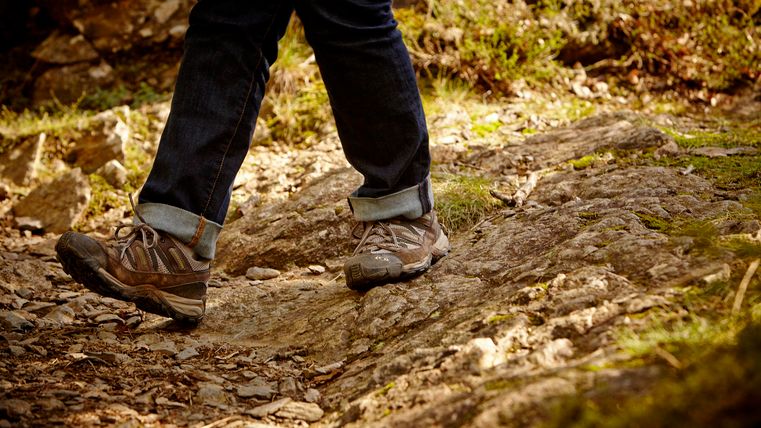 Eine Person wandert auf einem unebenen, steinigen Weg. Sie trägt robuste Wanderschuhe und ist in der Natur unterwegs.