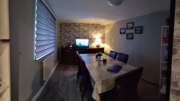 Ein gemütlicher Essbereich mit einem großen Tisch und Stühlen. An der Wand befindet sich ein Fernseher und es gibt eine warme Lichtquelle.