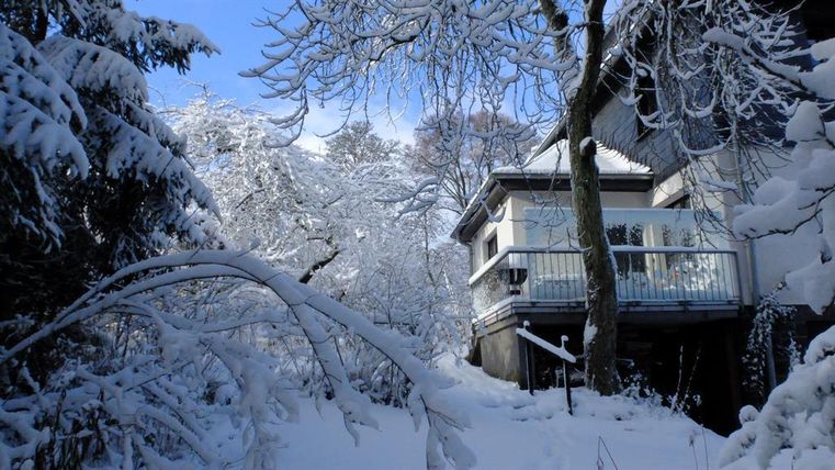 Ein verschneiter Garten mit einem modernen Haus in der Winterlandschaft. Die Bäume sind mit Schnee bedeckt und der Himmel ist klar und blau.