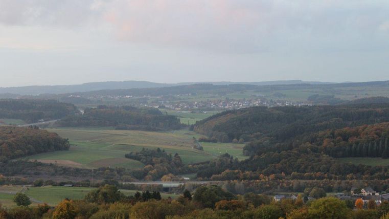 Eine ruhige Landschaft mit sanften Hügeln und weitläufigen Feldern. Im Hintergrund sind Wälder und ein kleiner Ort sichtbar.