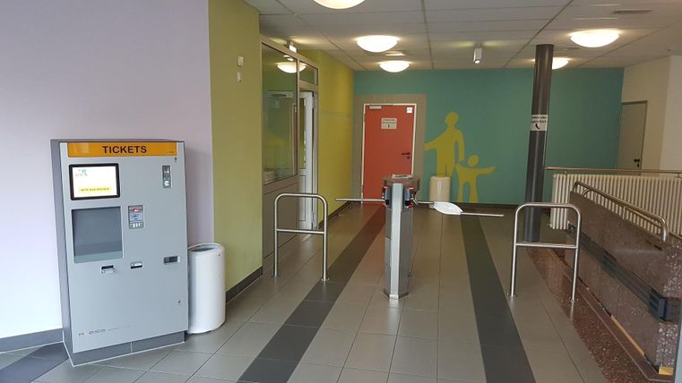 Ein moderner Eingangsbereich mit einem Ticketautomaten und automatischen Drehkreuzen. Die Wände sind in verschiedenen Farben gestrichen, was einen einladenden Eindruck vermittelt.