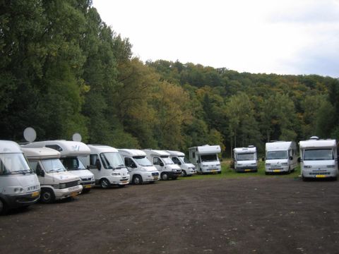 Eine Reihe von Wohnmobilen steht auf einem Parkplatz, umgeben von Bäumen. Der Himmel ist bewölkt und die Landschaft wirkt ruhig und idyllisch.
