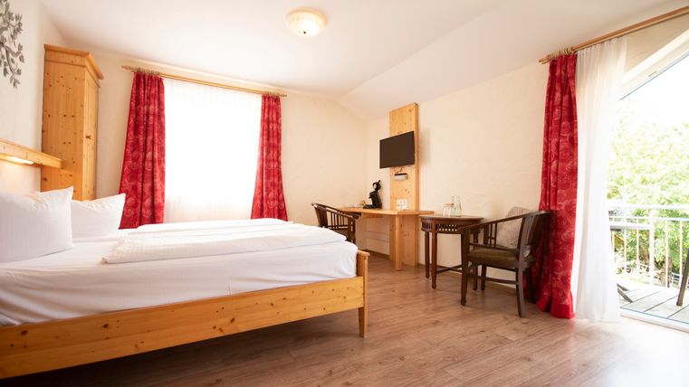 Ein helles Zimmer mit einem Doppelbett, einem Tisch und Stühlen. Große Fenster sorgen für viel Tageslicht und einen Blick nach draußen.
