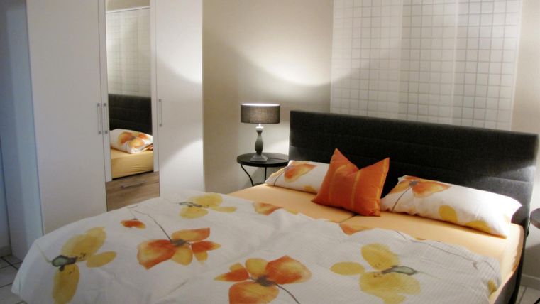 Ein modernes Schlafzimmer mit einem großen Doppelbett und bunten Blumendekoren. Im Hintergrund gibt es einen Spiegel und eine Lampe auf einem Nachttisch.