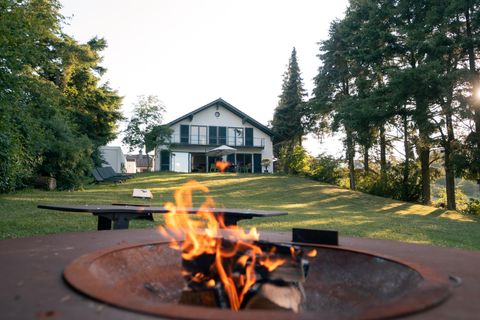 Ein schönes Haus umgeben von Bäumen und grünem Rasen. Im Vordergrund brennt ein Feuer in einem Grill.