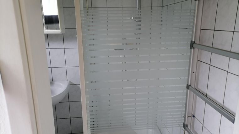 Ein modernes Badezimmer mit einer Duschkabine und gefliesten Wänden. Im Hintergrund befindet sich ein kleines Waschbecken und ein Spiegel.