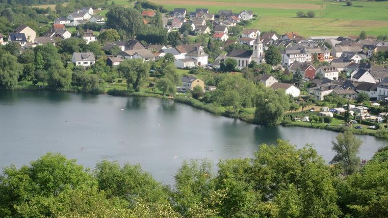 Eine malerische Landschaft mit einem ruhigen See und umliegenden Häusern. Grüne Bäume und Wiesen vervollständigen die idyllische Szene.