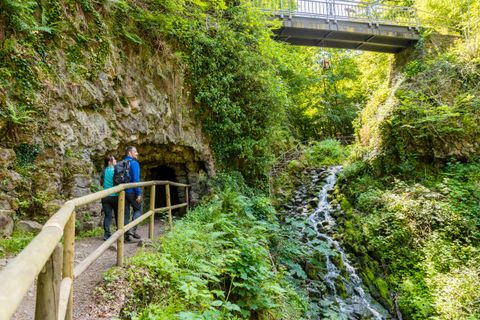 Zwei Personen stehen vor einer Höhle in einer grünen, bewaldeten Umgebung mit einem kleinen Wasserfall und einer Brücke darüber.