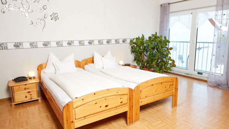 Ein gemütliches Schlafzimmer mit zwei Einzelbetten aus Holz. Es gibt ein Nachtkästchen, eine Pflanze und große Fenster mit Blick nach draußen.