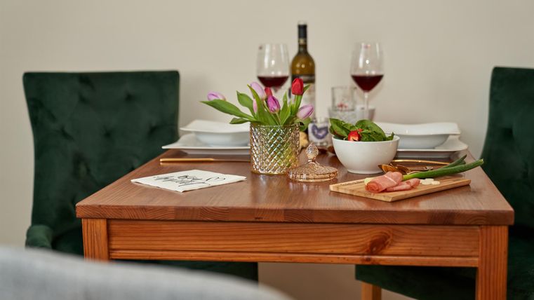 Ein schön gedeckter Tisch mit rotem Wein und frischen Zutaten. Im Hintergrund stehen grüne Stühle und eine dekorative Vase.