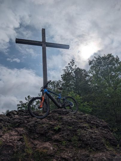 Ein Fahrrad steht auf einem Felsen vor einem Holzkreuz. Der Himmel ist bewölkt und es sind Bäume im Hintergrund zu sehen.