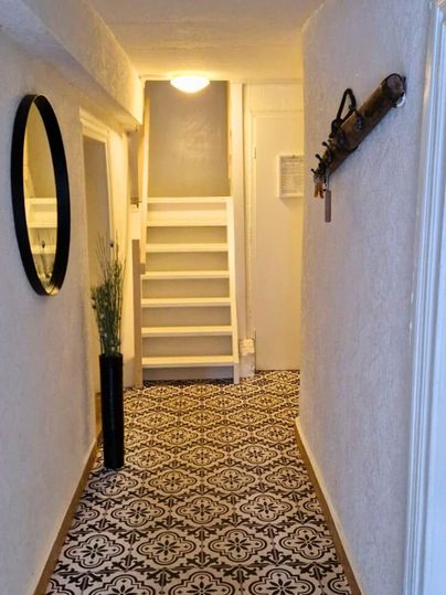 Ein heller Flur mit einem bunten Musterboden und einer Treppe am Ende. An der Wand hängt ein Spiegel und eine dekorative Waffe.