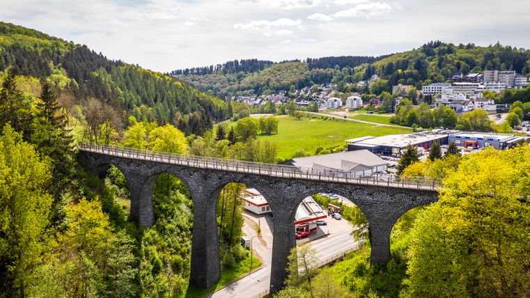 Luftaufnahme des Daun Viadukts auf dem Maare-Mosel-Radweg, umgeben von grünen Wäldern und einer Stadt im Hintergrund.