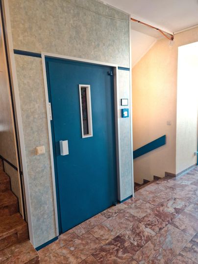 Ein Flur mit einem blauen Aufzug und einer Treppe. Die Wände sind hell und die Böden sind mit einem marmorierten Muster versehen.