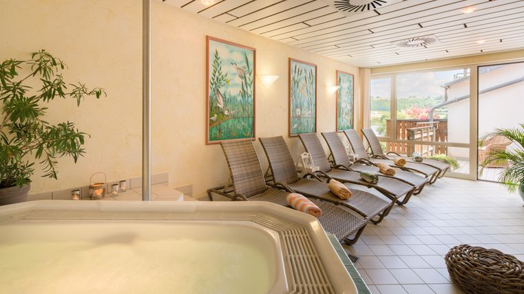 Ein entspannender Wellnessbereich mit einem Whirlpool und mehreren Liegen. Große Fenster bieten einen Blick nach draußen auf die grüne Umgebung.