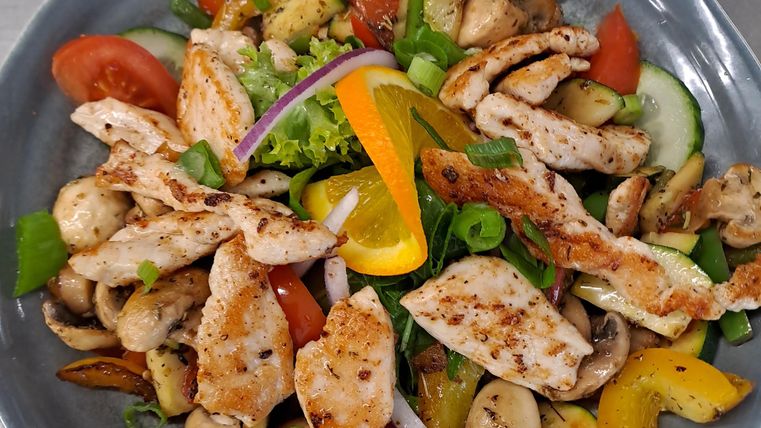 Ein bunter Salat mit gegrilltem Hähnchen, frischem Gemüse und Kräutern. Die Mischung umfasst Tomaten, Paprika, Gurken und Champignons.