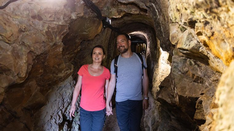Ein Paar geht Hand in Hand durch einen schmalen, beleuchteten Tunnel aus Fels. Die Atmosphäre wirkt abenteuerlich und interessant.