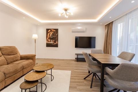 Ein moderner Wohnraum mit einer bequemen Couch, einem Tisch und einem Fernseher. Großfenster sorgen für viel Tageslicht und eine gemütliche Atmosphäre.