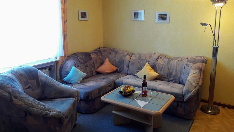 Ein gemütliches Wohnzimmer mit einer großen Couch und einem bequemen Sessel. Auf dem Tisch stehen Obst und eine Flasche Wein, während das Licht sanft durch das Fenster fällt.