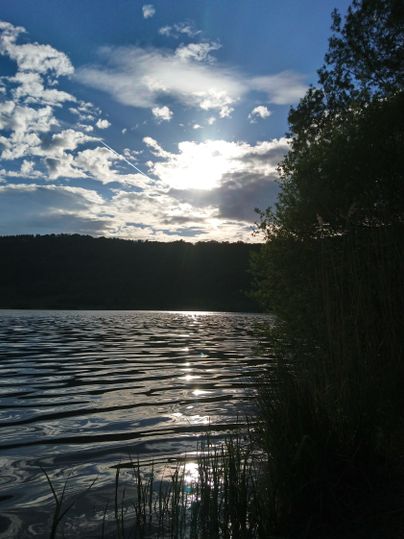 Ein ruhiger See bei Sonnenuntergang, umgeben von Bäumen und einem klaren Himmel. Die Wasseroberfläche spiegelt die Wolken und das Licht der Sonne wider.