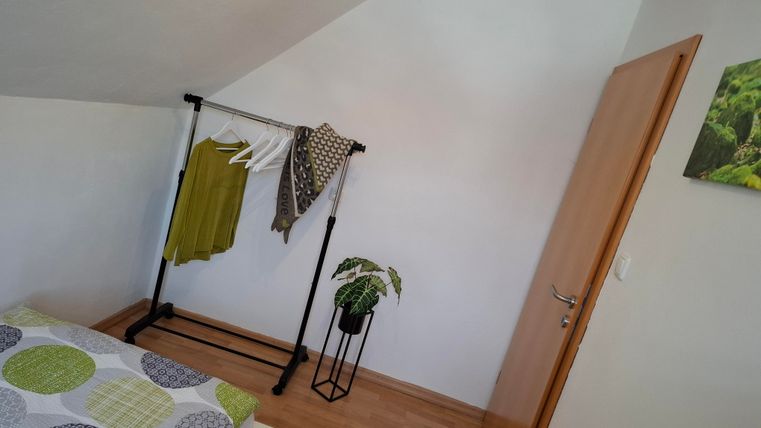 Ein helles Zimmer mit einem Bett und einem Kleiderständer. An der Wand hängt ein grünes Pflanzenbild, und es gibt einen kleinen Topf mit einer Pflanze.