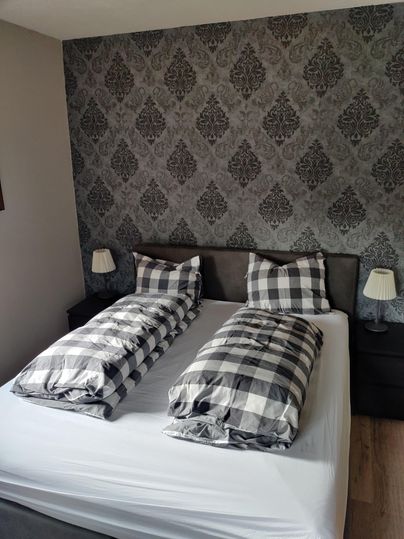 Ein modernes Schlafzimmer mit einem Doppelbett und einem stilvollen, gemusterten Hintergrund. Die Bettdecken sind in einem karierten Design und neben dem Bett stehen kleine Lampen.