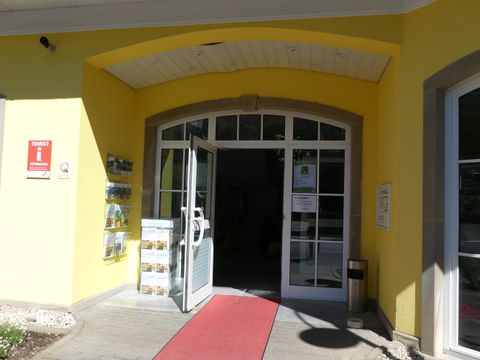 Der Eingang zu einem hellgelben Gebäude mit einer offenen Glastür. Ein roter Teppich führt hinein, und einige Informationsbroschüren sind daneben angebracht.