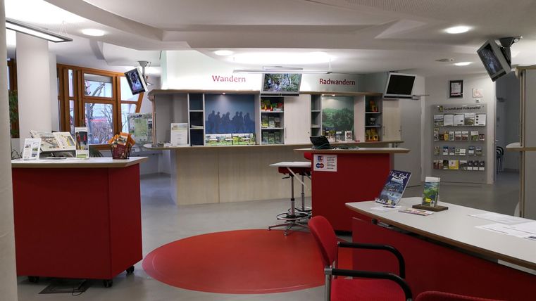 Ein modernes Informationszentrum mit einem Empfangstresen und roten Stühlen. Auf Regalen sind verschiedene Informationsmaterialien zu Wandern und Freizeitangeboten ausgelegt.