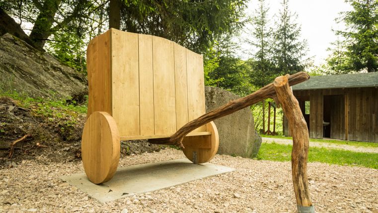 Wooden sculpture of a wagon on a forest path with trees in the background.