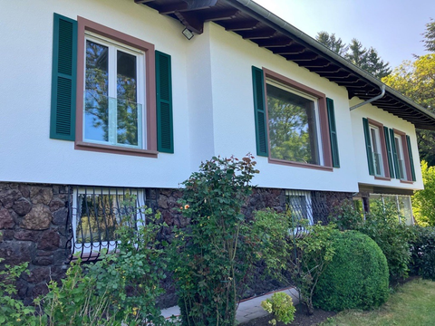 A beautiful house with white walls and green shutters. The garden is well-maintained and has many plants.
