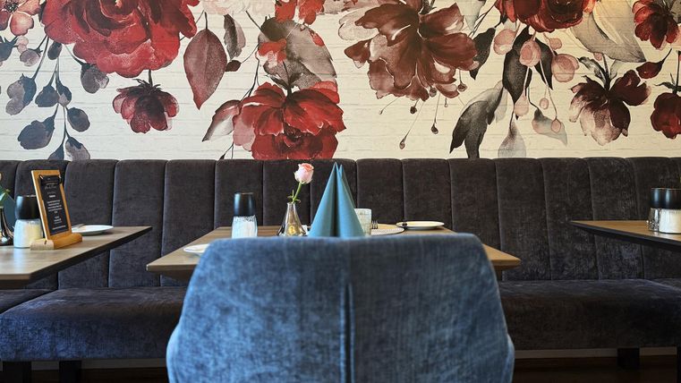 Ein modernes Restaurant mit einer eleganten Wandbemalung aus roten Blumen. Die Einrichtung ist gemütlich mit blauen Sitzbänken und stilvollen Tischdekoren.