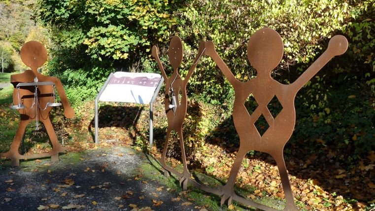 Drei große, metallene Figuren stehen an einem Weg, umgeben von buntem Herbstlaub. Eine Informationsstele befindet sich zwischen den Figuren.