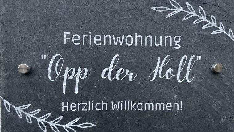Ein Schild für eine Ferienwohnung mit dem Namen "Opp der Holl". Es heißt "Herzlich Willkommen!" und hat eine dekorative Schrift und Pflanzenmotive.