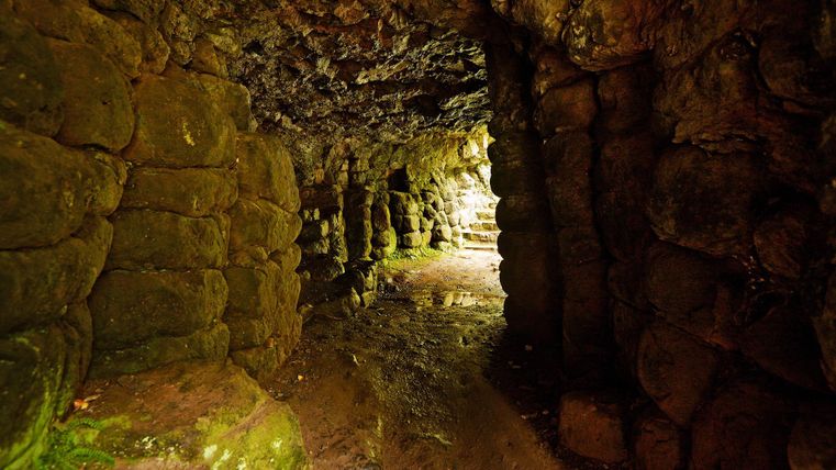 Ein schmaler, feuchter Gang in einer Höhle mit einer unregelmäßigen Steinmauer. Das Licht am Ende des Gangs sorgt für eine mystische Atmosphäre.