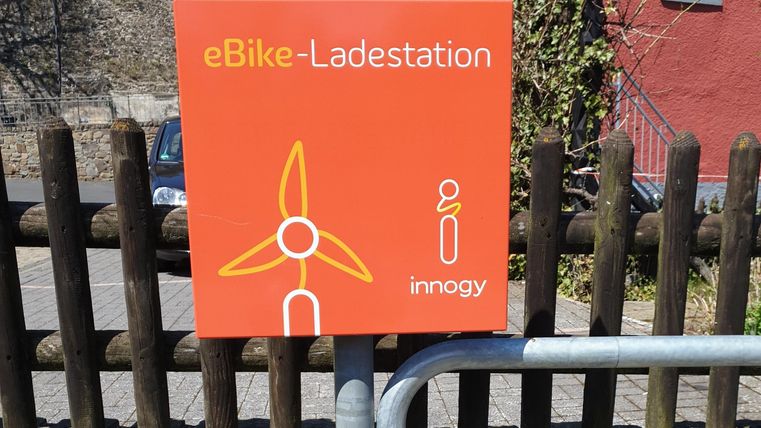 Eine eBike-Ladestation mit einem orangefarbenen Schild und einem Windrad-Symbol. Im Hintergrund ist ein Holzlattenzaun und ein rotes Gebäude zu sehen.