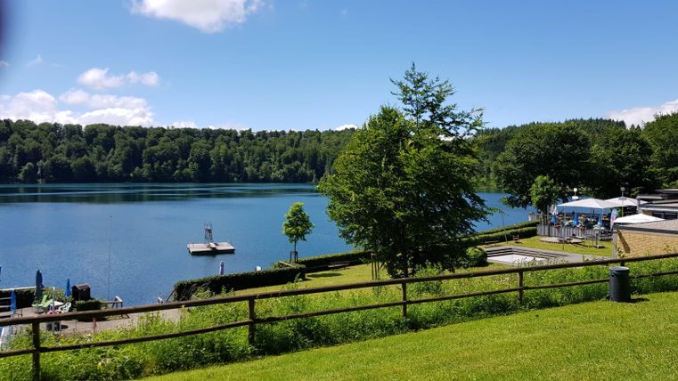 Ein ruhiger See umgeben von grünen Bäumen und Wiesen. Im Hintergrund sind ein Steg und einige Liegestühle zu sehen.