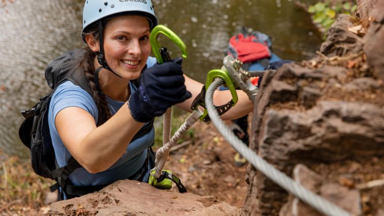 Eine Frau klettert an einer Felswand und lächelt dabei. Im Hintergrund sind Kletterausrüstungen und ein Fluss zu sehen.