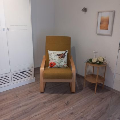 A cozy armchair with a colorful cushion is in a bright room. Next to it, there is a small table with flowers and a decoration.