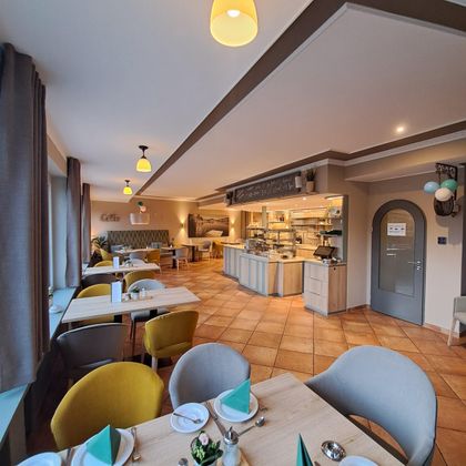 Ein modernes Restaurant mit hellen Wänden und gemütlichen Tischen. Es gibt Bänke und Stühle in verschiedenen Farben sowie eine offene Küche.