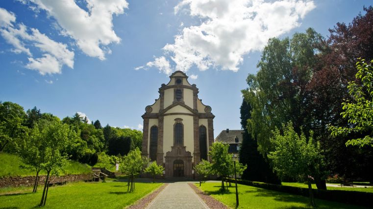 Eine beeindruckende Kirche umgeben von grüner Landschaft und Bäumen. Der Himmel ist klar mit einigen Wolken.