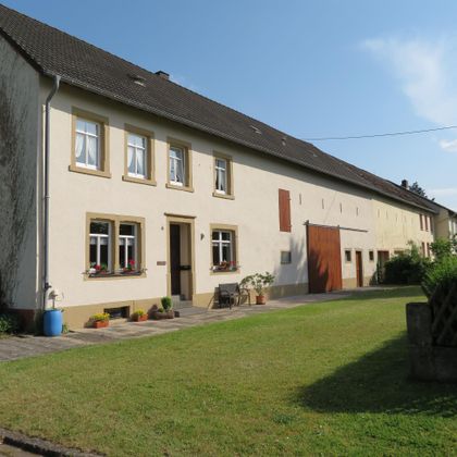 Ein großes, helles Haus mit mehreren Fenstern und einem grünen Vorgarten. Der Himmel ist blau und klar.