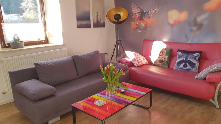 Ein modernes Wohnzimmer mit einem grauen Sofa und einem roten Sofa. Auf dem Tisch stehen frische Blumen und es gibt dekorative Kissen mit Motiven.