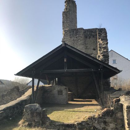 <p>Im Hintergrund sieht man eine hochaufragende etwa 5 Meter breite Mauer. An deren rechter Seite setzt eine im rechten Winkel noch höhere Wand aus Bruchsteinen an.<br/>Vor der Wand hat man eine Holzkonstruktion als Dach errichtet, die an der hinteren Wand abschließt. Sie ist an beiden Seiten und nach vorne offen. Ganz im Vordergrund sieht man einige Bauerreste, die teilweise mit Gras bewachsen sind. In den  Flächen zwischen den Mauerresten hat sich ebenfalls Gras ausgebreitet.</p>
