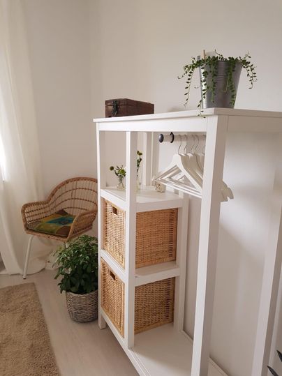 Ein heller Raum mit einem weißen Regal, das Körbe und Kleiderbügel enthält. In der Ecke steht ein gemütlicher Korbsessel und einige Pflanzen sorgen für eine frische Atmosphäre.
