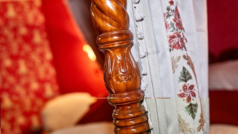 Ein dekoratives Kopfteil aus Holz mit einer gedrehten Säule. Im Hintergrund sind rote Wände und ein Bett zu sehen.