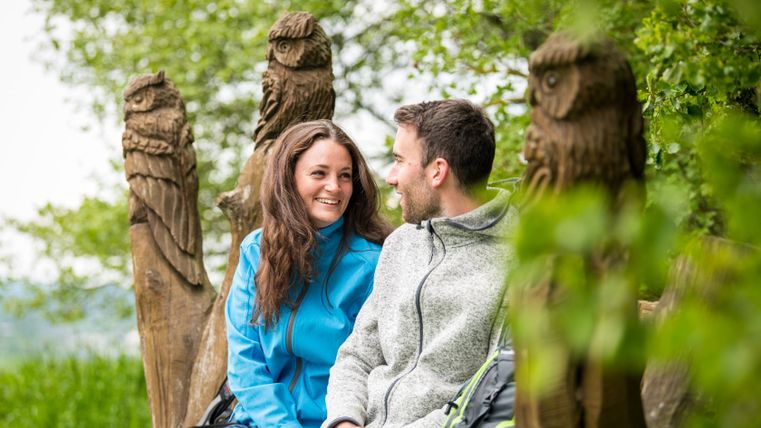 A couple sits on a bench with owl carvings outside.